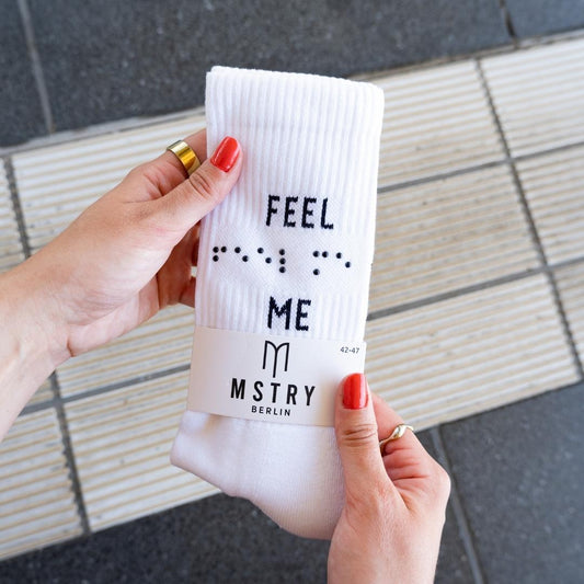 Zwei Hände halten die neue FEEL ME Socke über einen Leitstreifen. Auf der weißen Socke steht das Statement FEEL ME. Zwischen den zwei Wörtern ist die Botschaft in Braille mit Kuppen aufgepresst.